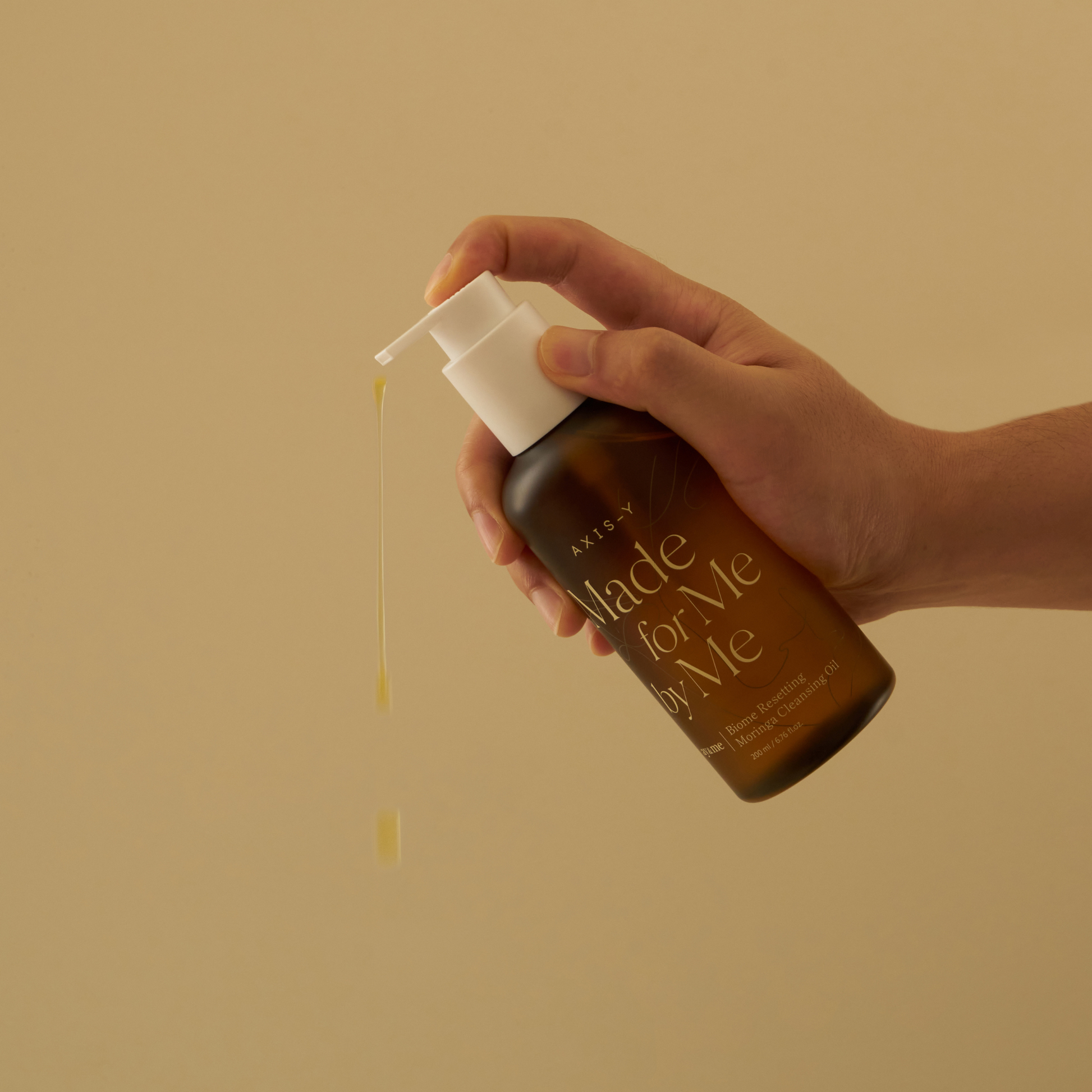 AgelessBeauty3000.com  AXIS-Y  |  Biome Resetting Moringa Cleansing Oil  Hand holding a brown bottle with a pump, labeled 'made for Me by Me', against a beige background.
